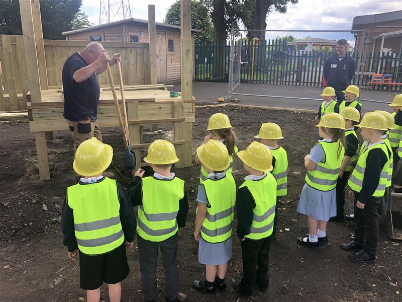 children in yellow hard hats and mini hi vis vests stand talking to an installer as he explains the construction process whilst they are having a playground built