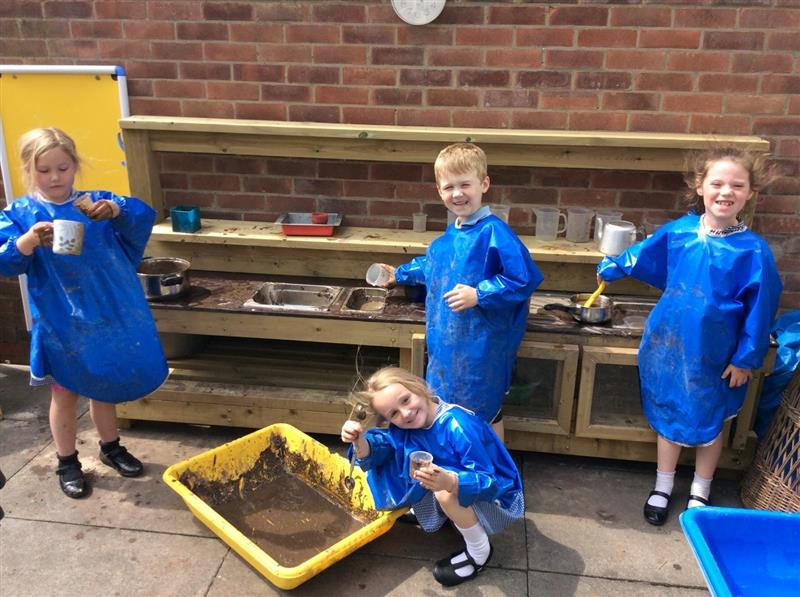 children in school uniform and blue waterproof aprons stand in front of a mud kitchen and play with sand and mud