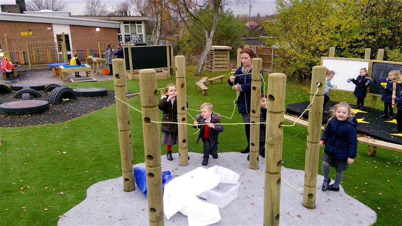 three children in navy winter coats and red school uniform stand with a teacher holding onto pieces of string that are ooped through den-making posts and tied neatly