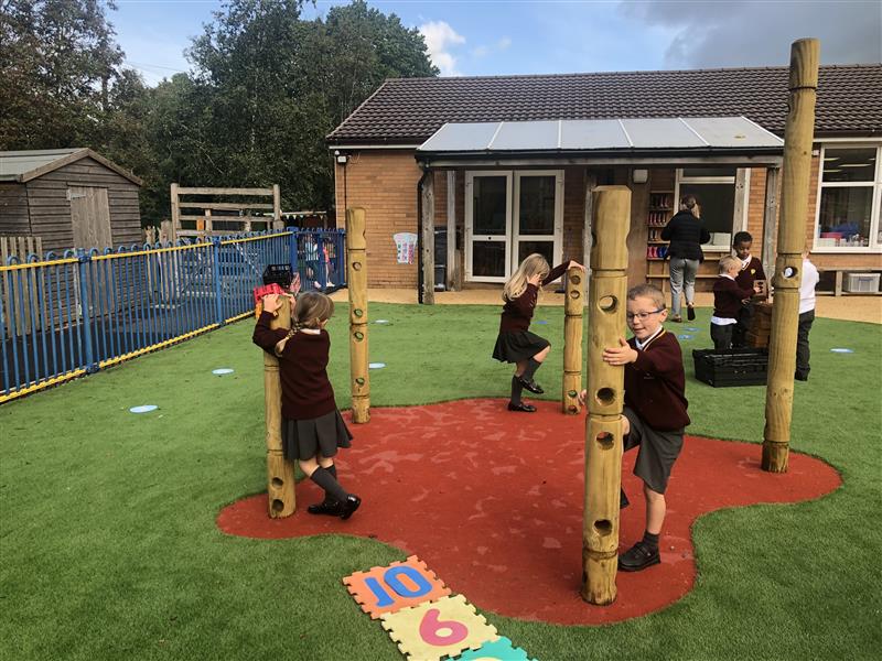 three pupils in burgundy school uniform stand on the red saferturf splodge and hang and swing around the den posts as they play