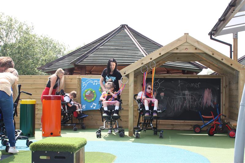 two chuldren in wheelchairs sit and enoy sensory products with the gaint playhouse in the background and the african drums and grass topped seats on the playground also