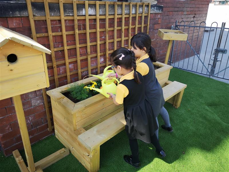 children kneel on the planter with benches and water the plants in the box with a yellow watering can