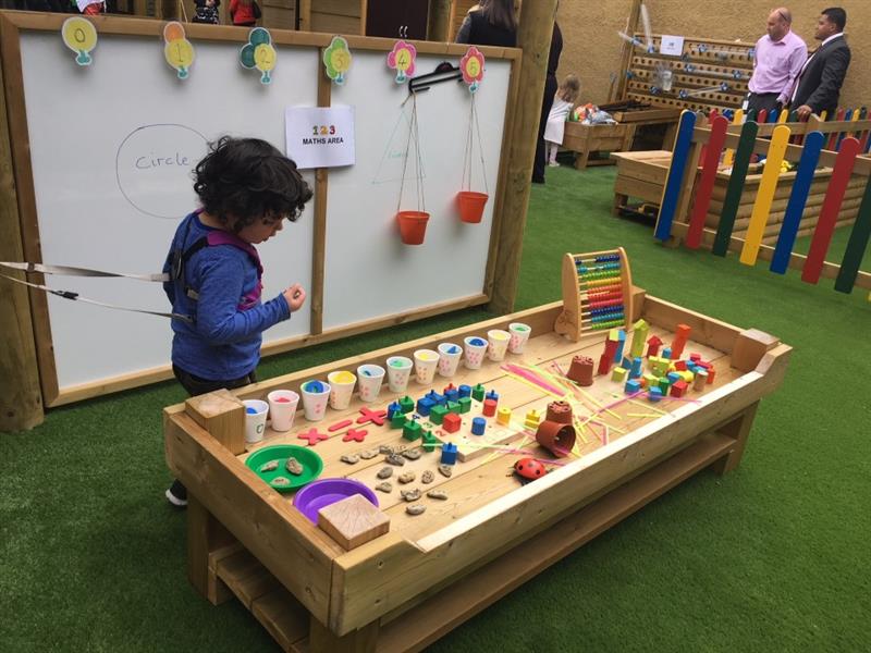 a little boy stands between a giant whitebard panel and a construction table, the table is covered with paint and crafting items 