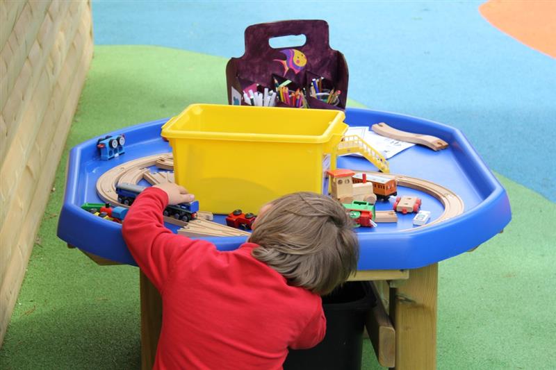 a child sits on the surfacing on the floor and plays at the blue tuff spot table with toys on it