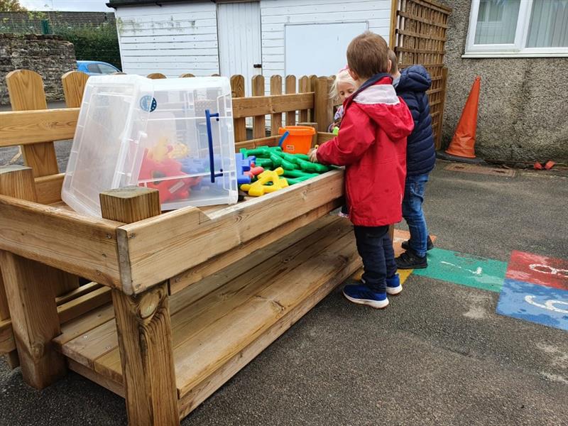 a little boy in a red coat stands assembling items at the construction table whilst taking to his friend in a navy coat