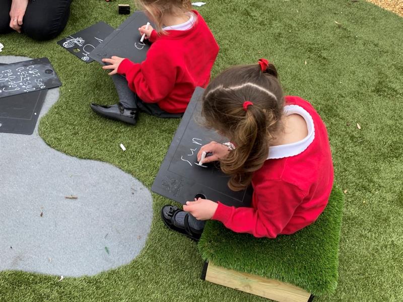 a little girl sits and writes on the scribble board on her knee whilst she sits on the green artificial grass