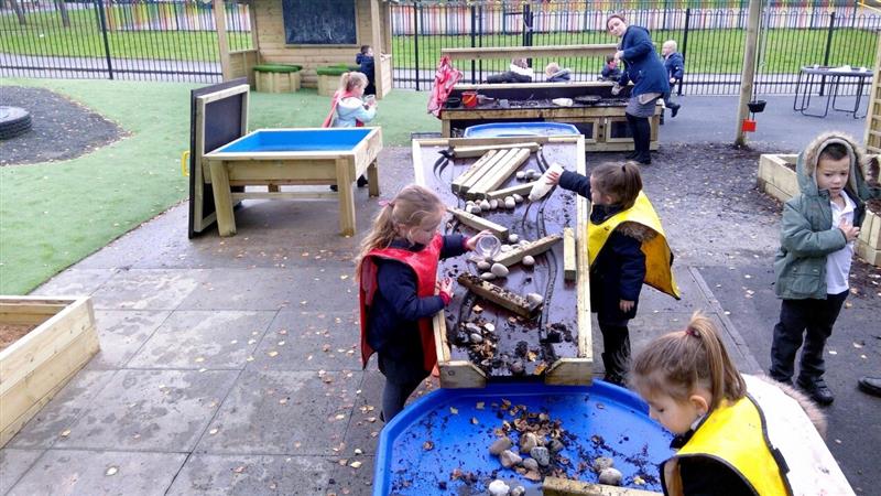 children wear read and yellow aprons as they play with the damming station that is covered in mud and water