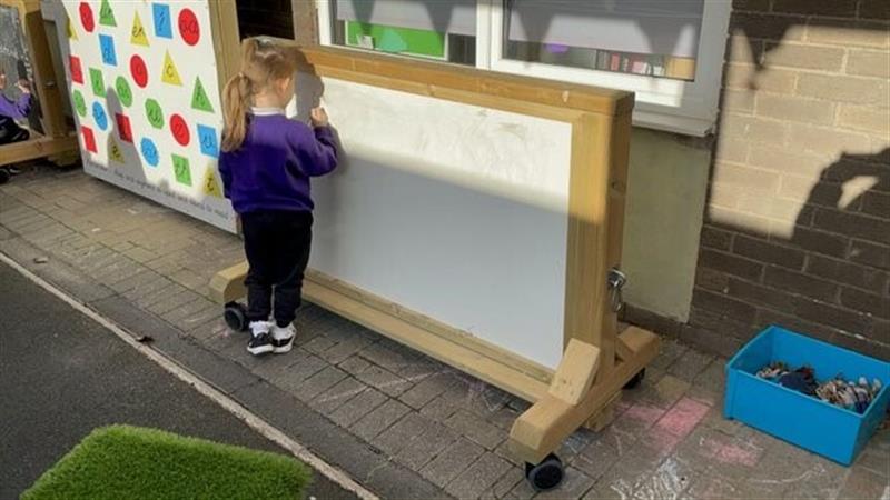 a little girl in purple school uniform stands in front of the whiteboard on wheels drawing