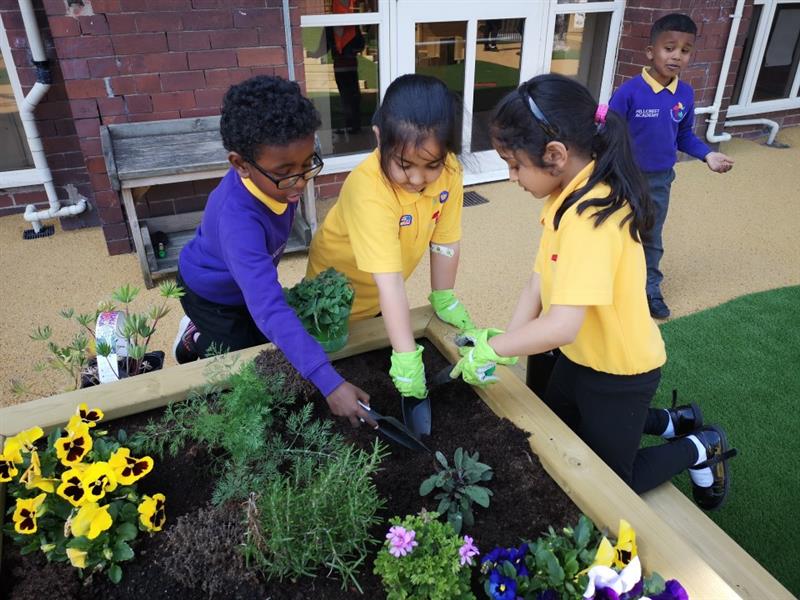 children stand around the planter with colourful plants inside and they dig at it with shovels and spades whilst wearing green plastic gloves