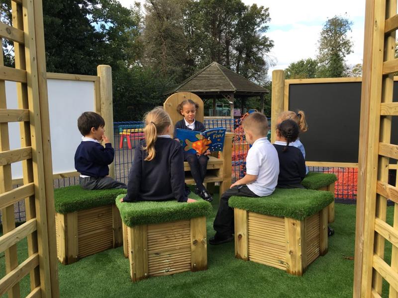 a child sits in the story telling chair reading a story to her classmates who sit on the artificial grass topped seats listening