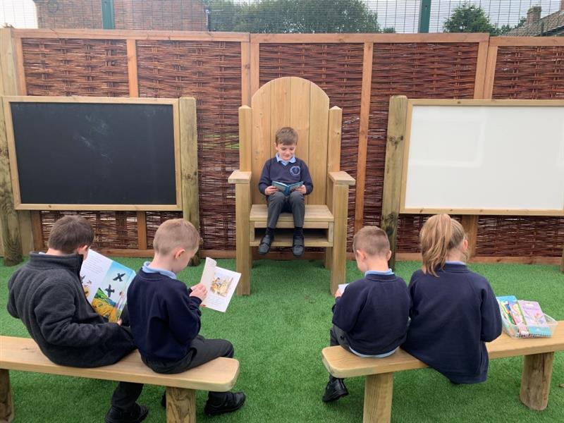 one child sits on the timber story telling chair reading a book whilst his classmates sit on the perch benches and read the storybooks