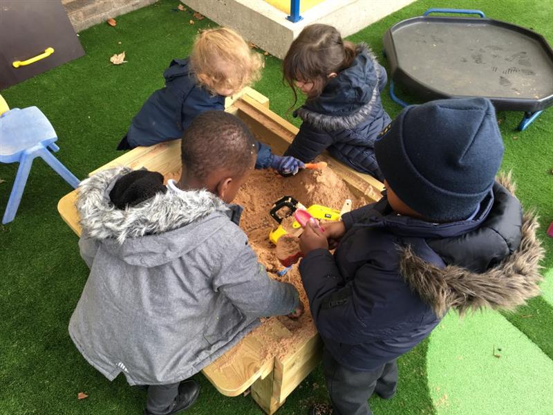 four children in hats and winter coats stand around a sand table and dig with their hands a little spades