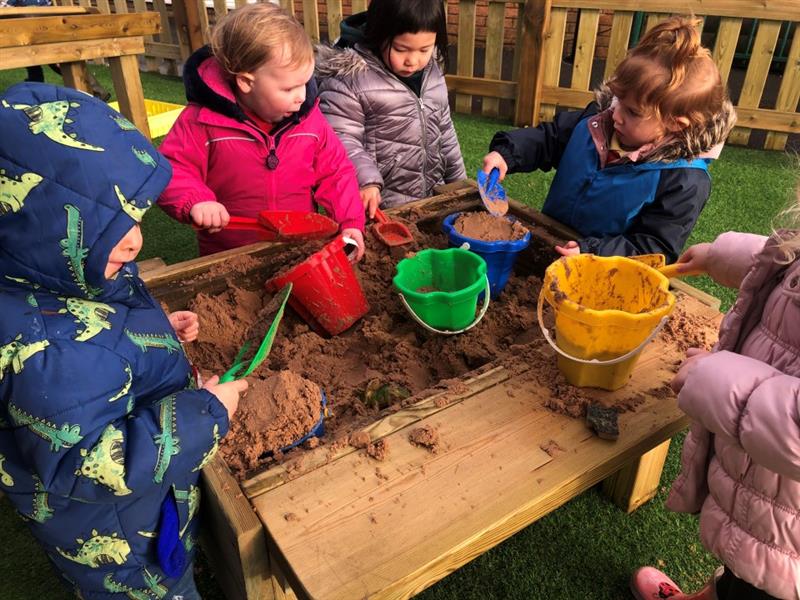children in winter coats sit around the sliding sand pit with buckets and spades and fill their buckets with sand