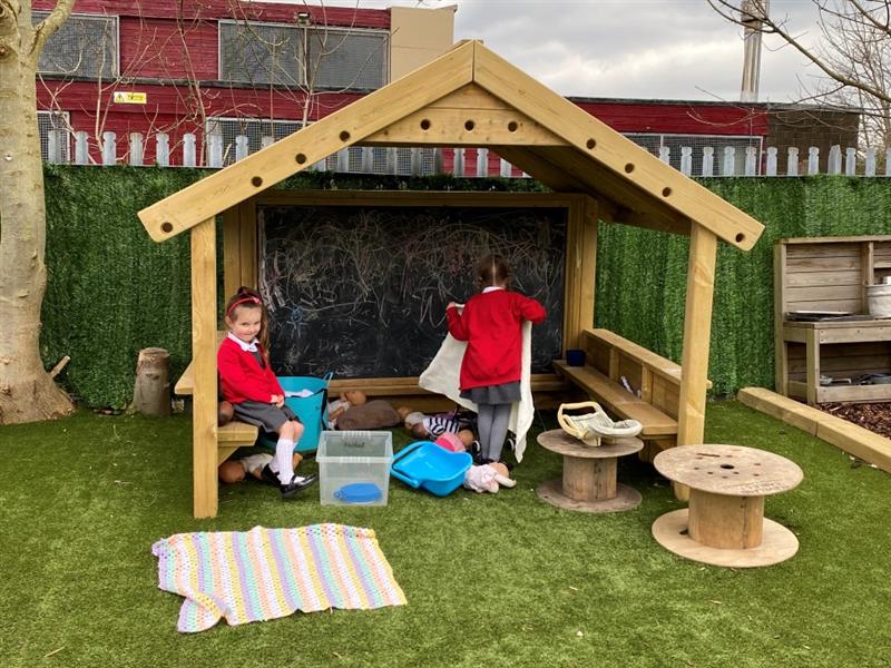 children in red school uniform stand within the playhouse with the chalkboard and the benches 