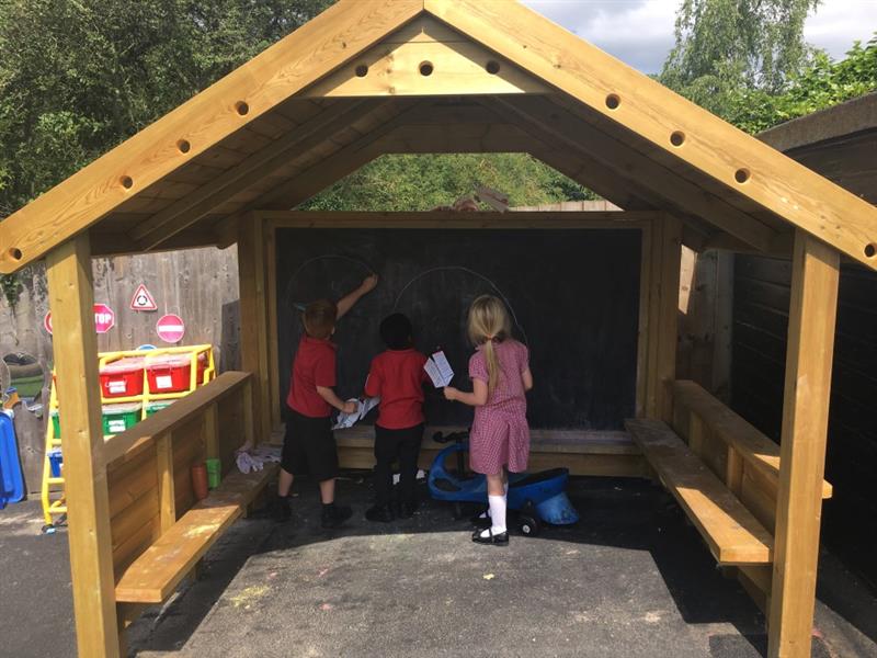 three children in red school unfiorm gather under the playhouse in front of the chalkboard and draw on the board with chalk