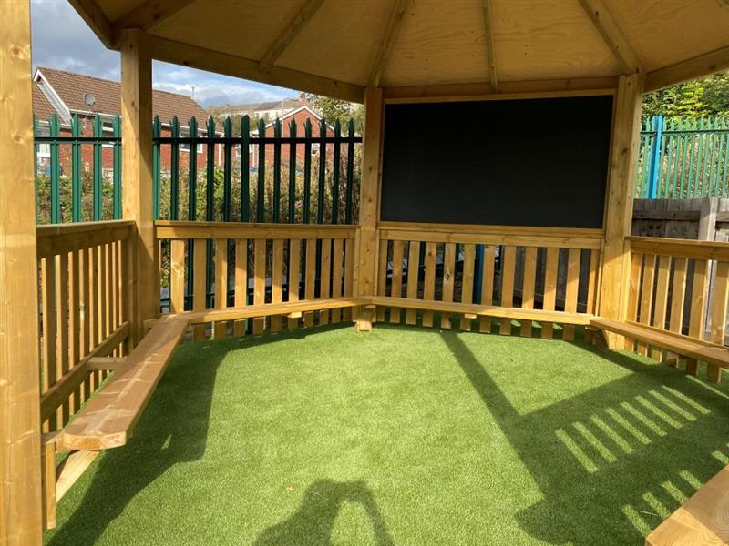the inside of the timber gazebo shows timber benches around the inside, a giant black chalkboard and artificial grass surfacing on the floor below
