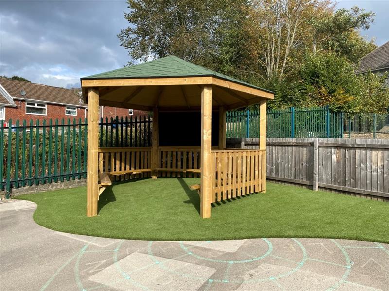 a timber gazebo with green roof and without a decked base on gree artificial grass surfacing with a giant black chalkboard and timber benches inside