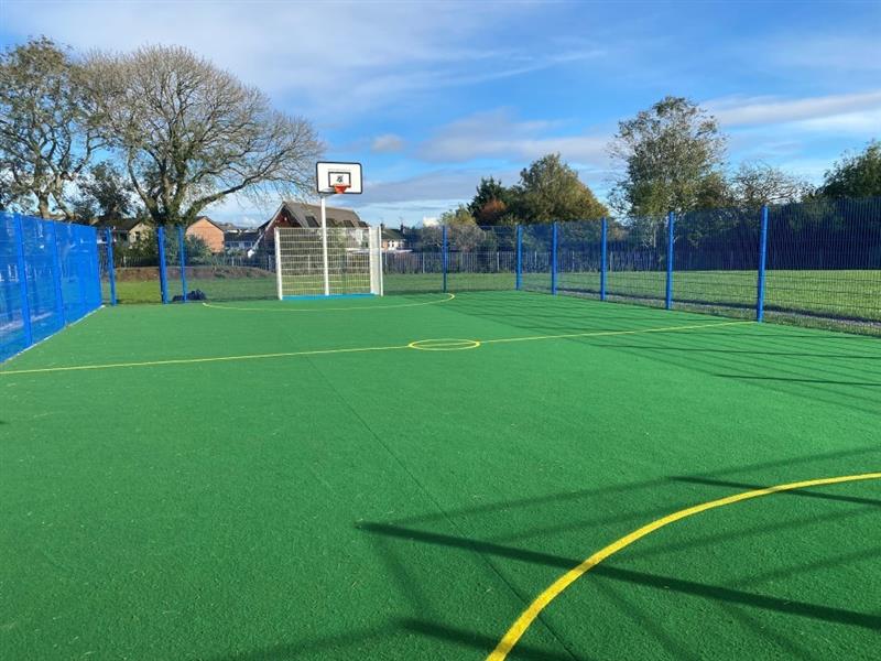 a view of the green and yellow muga surfacing with blue fences around and a basketball hoop