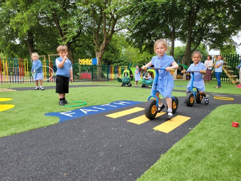 a little girl in a blue and white school dress scoots alogn the wetpour roadway with children running around on the artificial grass surfacing
