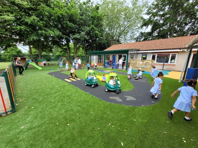 children drive around in plastic cars on the black wetpour roadway and green artificial grass surfacing 