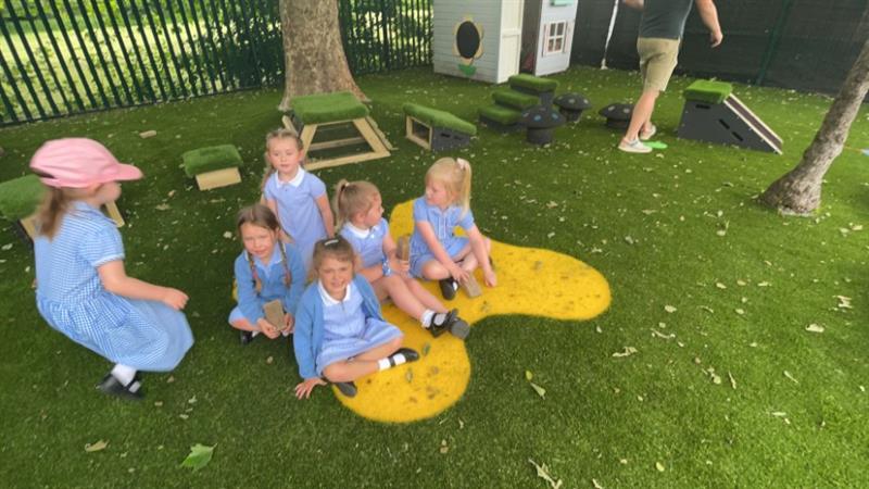 six children in blue school dresses sit on the green artificial grass with a yellow saferturf splodge  