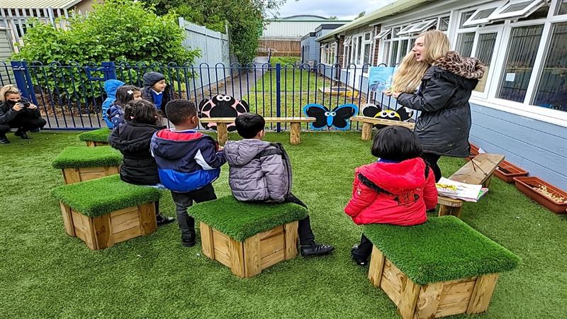 a teacher stands and reads a story to the children as they sit on the artificial grass topped seats listening and participating