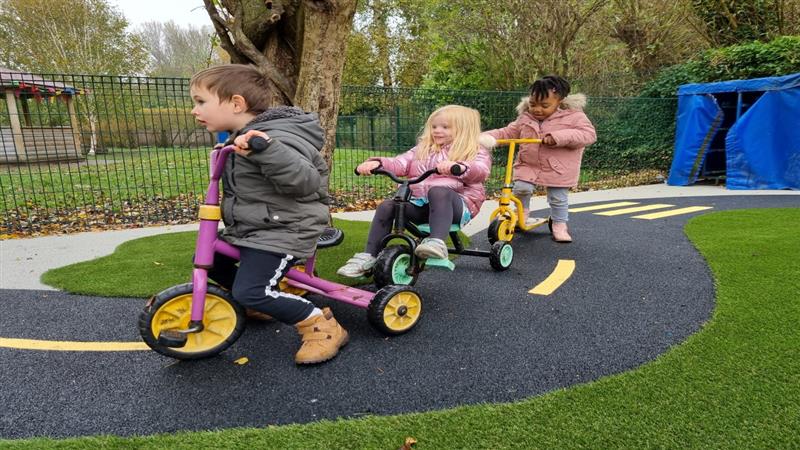 three children in winter coats scoot round on small scooters on the wetpour roadway