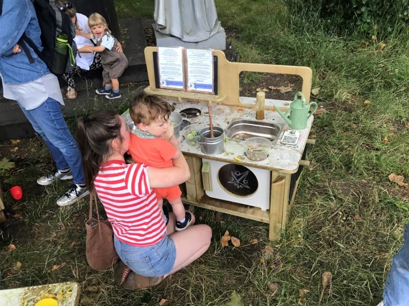 a mother holds her toddler up to the mud kitchen and helps him play with the materials in the mud kitchen