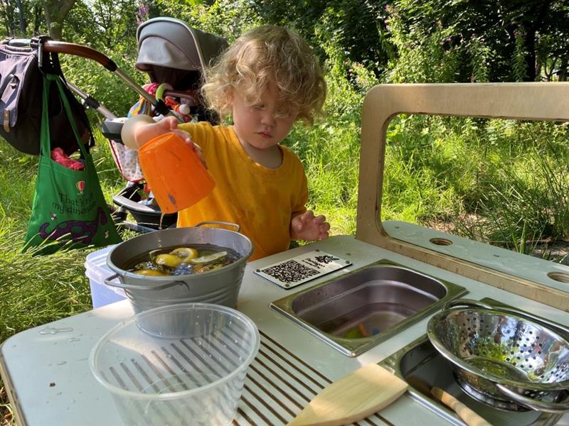 a child pour water in a bucket full of lemons, limes an other materials on the mud kitchen