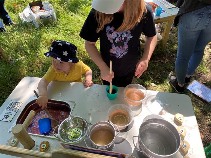 a child in a yellow top and black and white starry bucket hat mixes materials in the mud kitchen