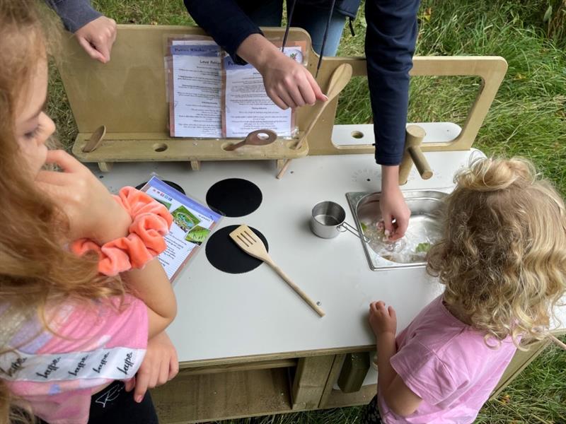 two little girls dressed in pink play with the mud kitchen whilst being supervised by an adult