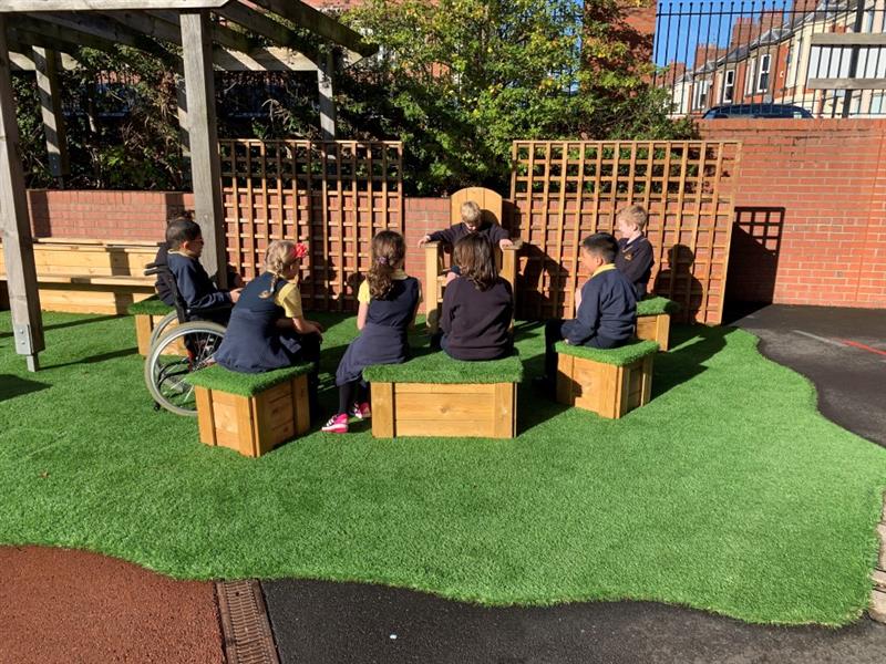 a little boys sits in the timber story telling chair and reads a story to his classmates who sit and listen carefully on the grass topped seats