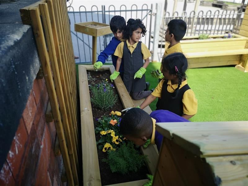 children stand and look at each other as the talk about what they are planting in the planter to their left