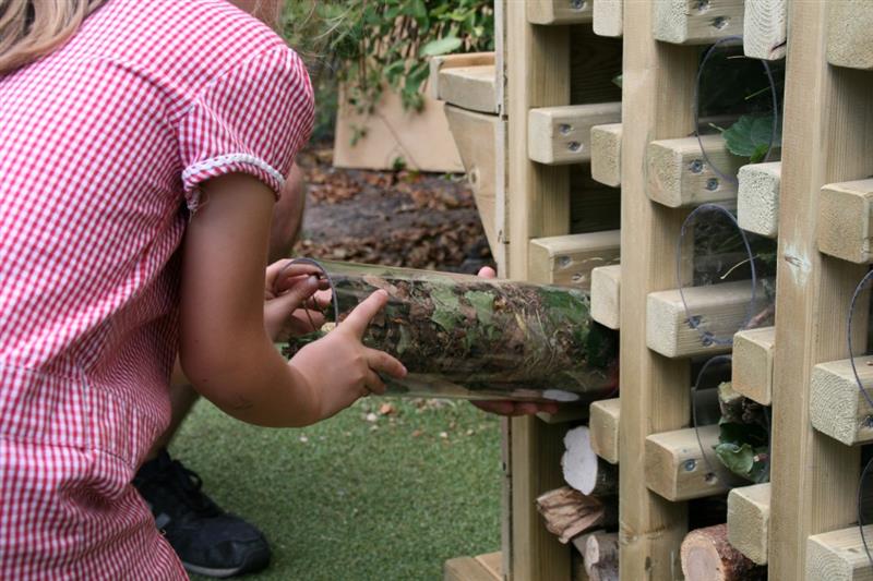 a child in a red and white dress kneels on the floor in front of the timber bug hotel and takes out one of the plastic tubes