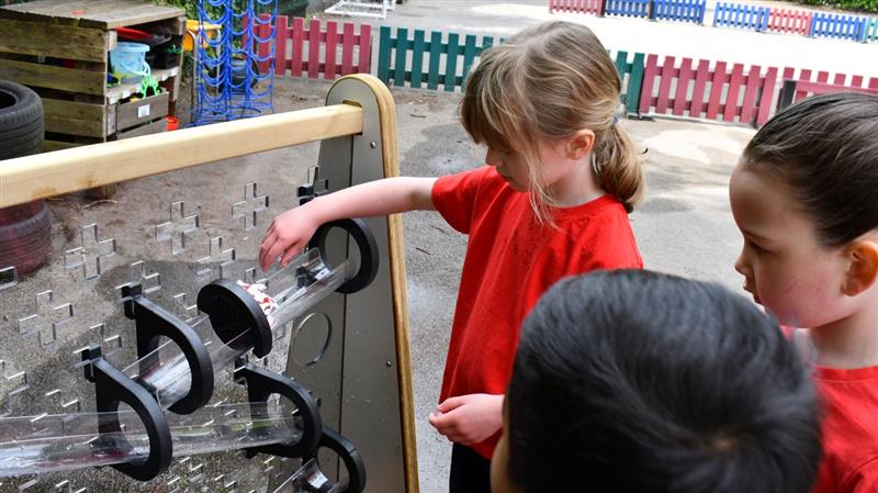 a child stands and pours water down a water channel stand on the water wall whilst her friends watch