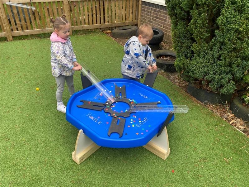 two children stand and send little plastic counters down the tube into the tuff spot table