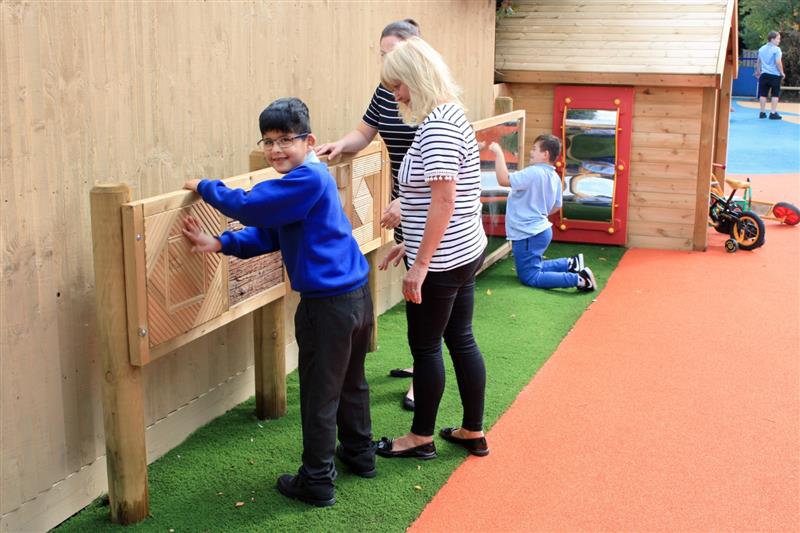 A child in blue school uniform using a sensory play panel on posts made up of different textured timbers, secured to two posts and installed onto artificial grass