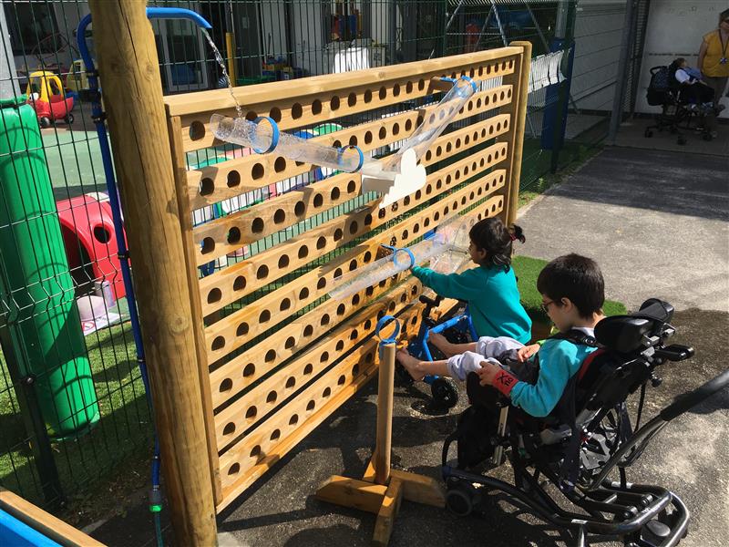Two children playing with pentagon plays water wall. One child in a wheelchair has their feet under the water whilst the second child using a walking aid lets the water splash on their hands