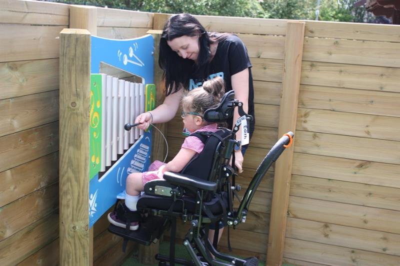 A child in a wheelchair sitting in front of outdoor musical play equipment whilst a teacher hits the silver chimes with a beater
