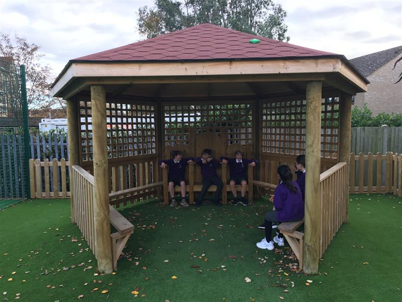 4 children sat inside of a timber outdoor classroom talking with one another