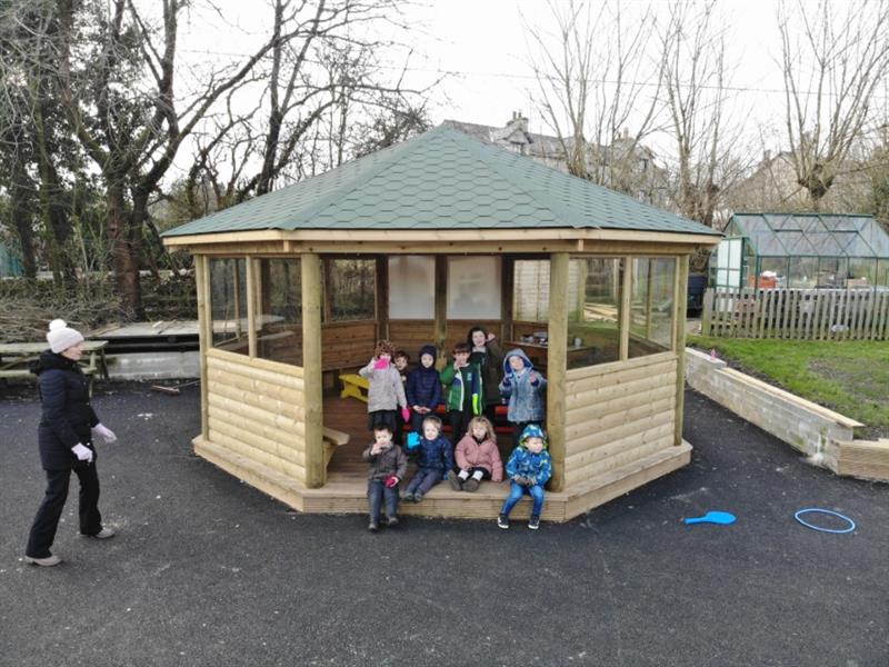 9 children in winter coats sat at the entrance of a decked outdoor gazebo waving at the camera