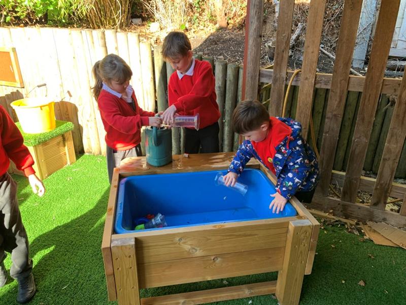 children lean over a water table with lid and they pour water between test tubes and watering can, they wear red school uniform and one wears a winter coat