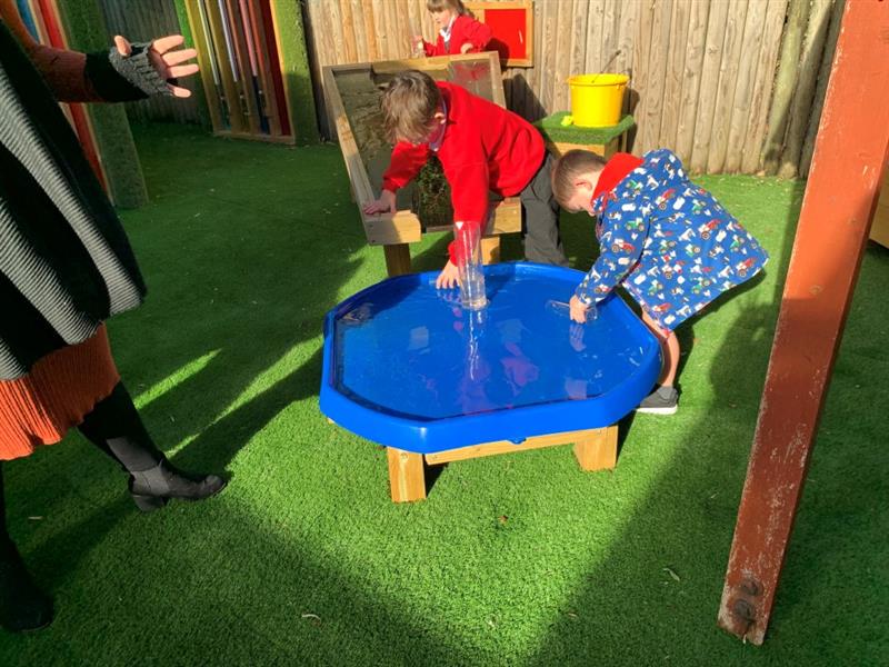 a child in red school uniform leans over the blue tuff spot table whilst a child in a blue patterned winter coat leans over the table too playing with the water