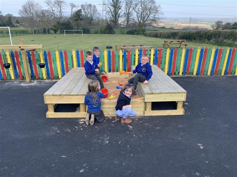 five children playing in a sandbox, four children are sat on the edges of the sand box 