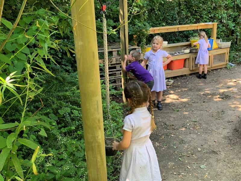 children play with both the mud kitchen and the water pulley, they are wearing purple and white school uniforms