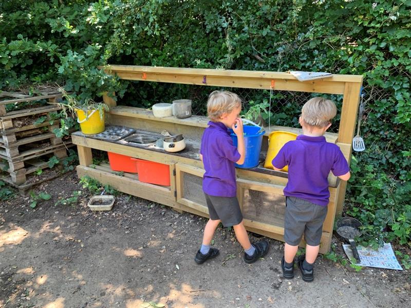 two young boys in purple school uniform play with the mud kitchen using a blue and a yellow bucket respectively, there is a bush in the background behind them
