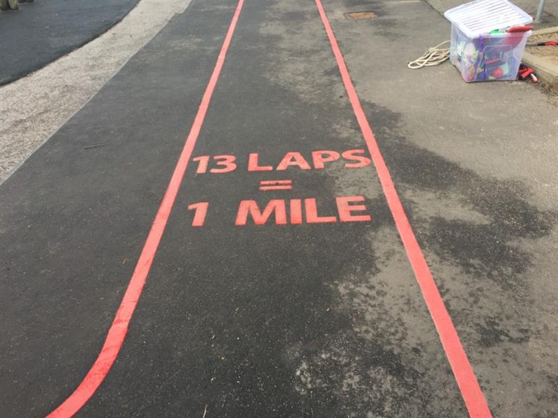 Red line playground markings adjacent to one another to create a running route on tarmac with the words 13 laps equals 1 mile in the center