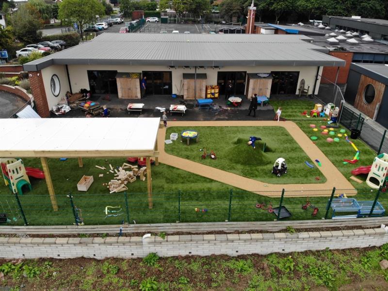 artificial grass surfacing on the playground viewed from a birdseye view angle, it shows the top of the timber canopy roof as well as the beige wetpour roadway and various nursery toys