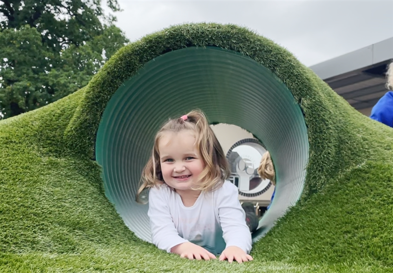 a little girl lies in the bespoke playground mound with tunnel smiling at the camera in front of her