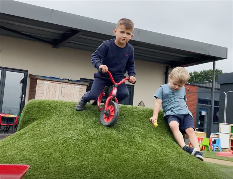 a children in dark navy clothes on a red scooter is pictured riding over the playground mound whilst his friend stands next to him